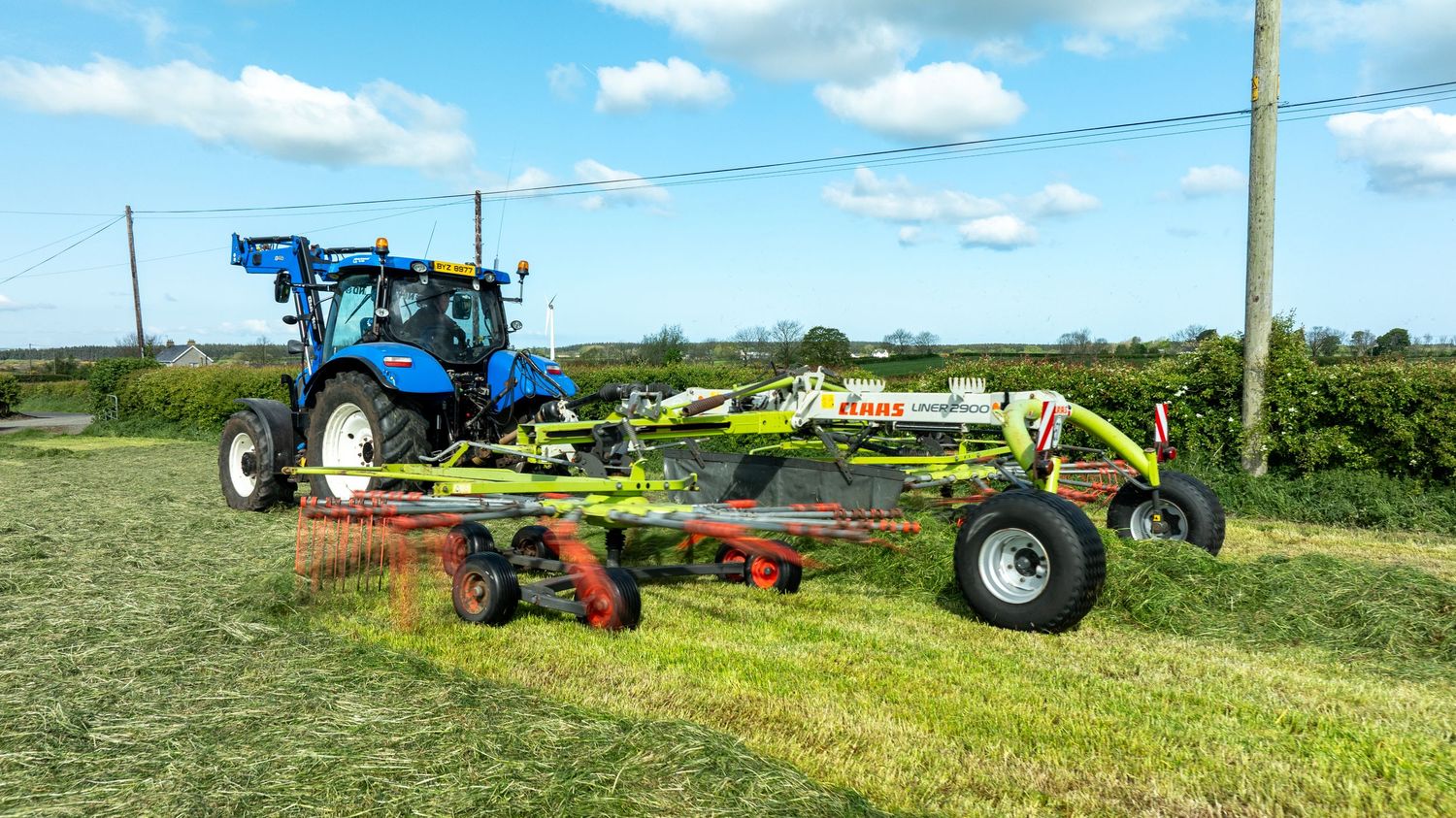 Blue tractor pulling a hay raking implement across a grassy field on a sunny summer day with blue skies.