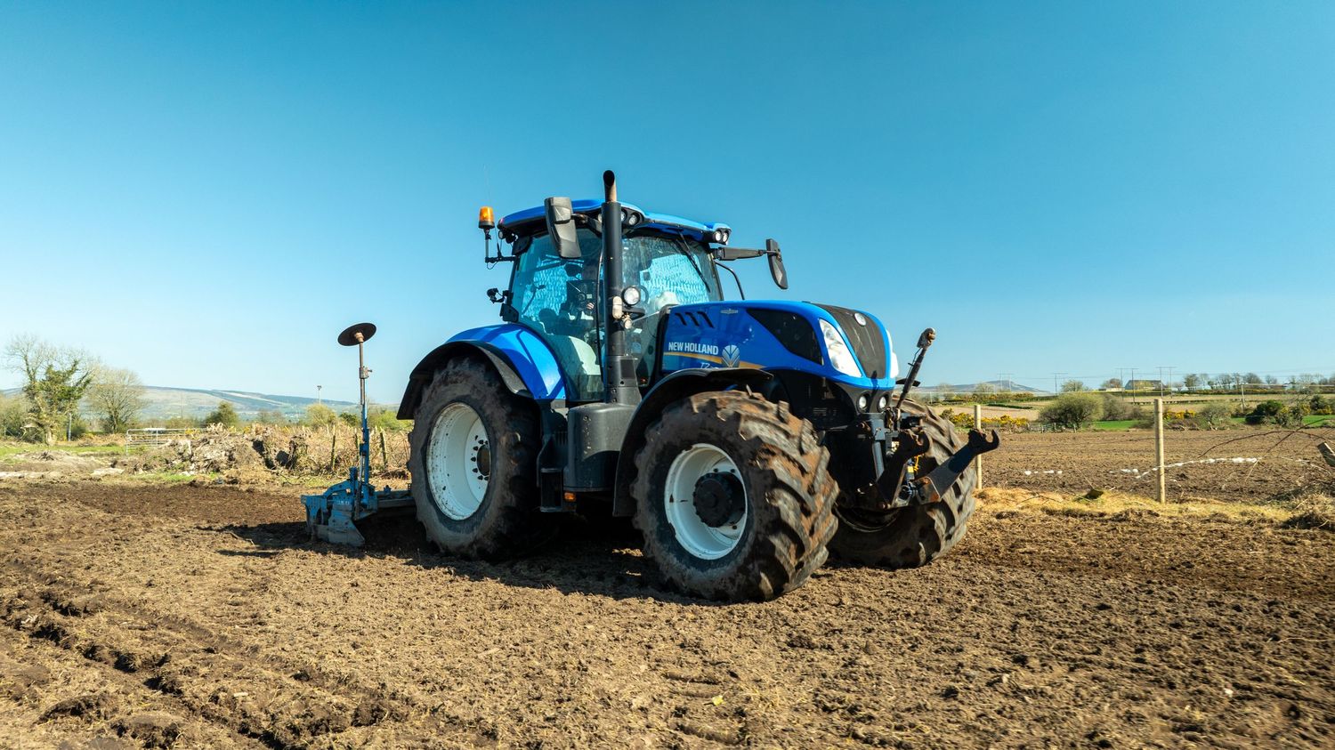 A bright blue New Holland tractor plowing a field against a clear sky in an agricultural setting.