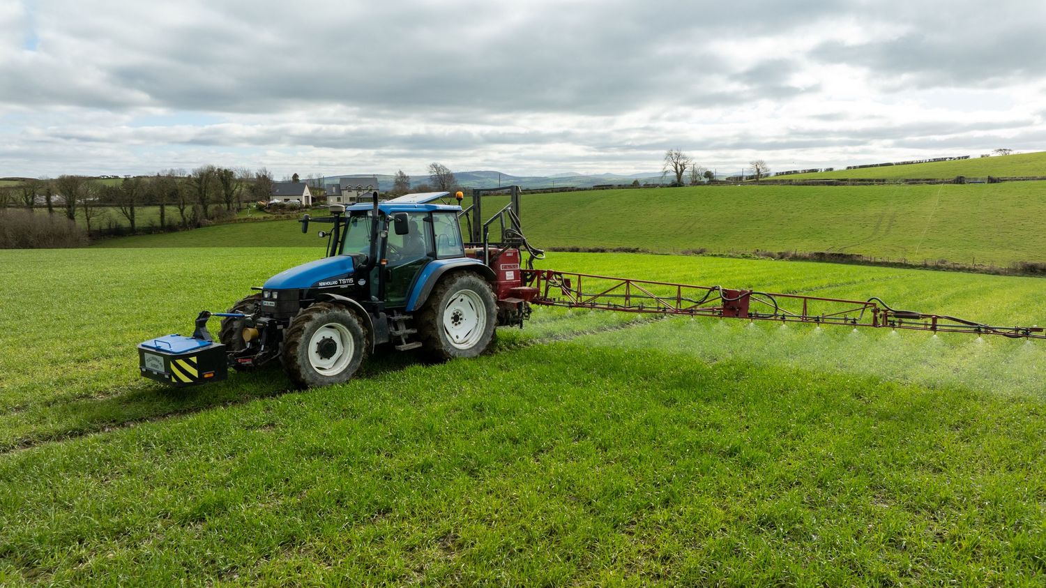 Blue tractor spraying fertilizer across green farmland under cloudy skies.