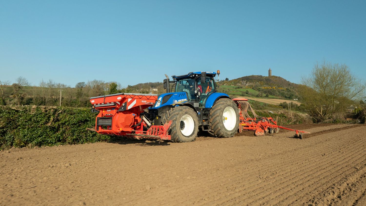 A blue New Holland tractor pulling red farming equipment works to prepare agricultural soil on a sunny day.