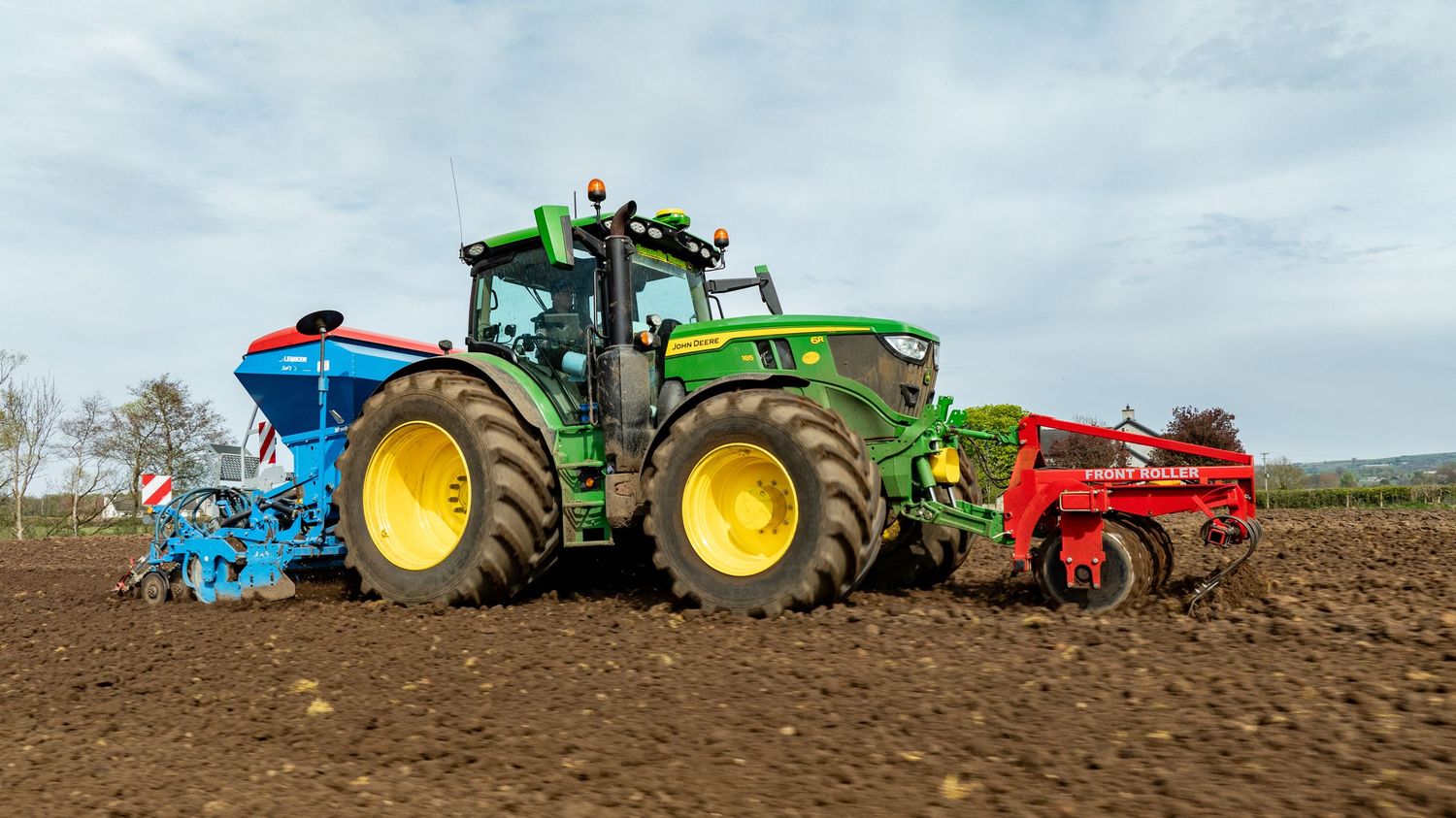 Green John Deere tractor with blue and red farming implements working in a freshly tilled field.
