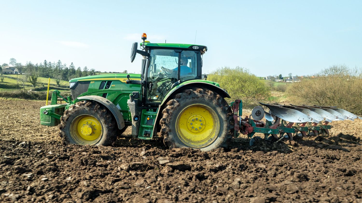 A green John Deere tractor plows dark soil in a rural field with blue cultivation equipment attached.