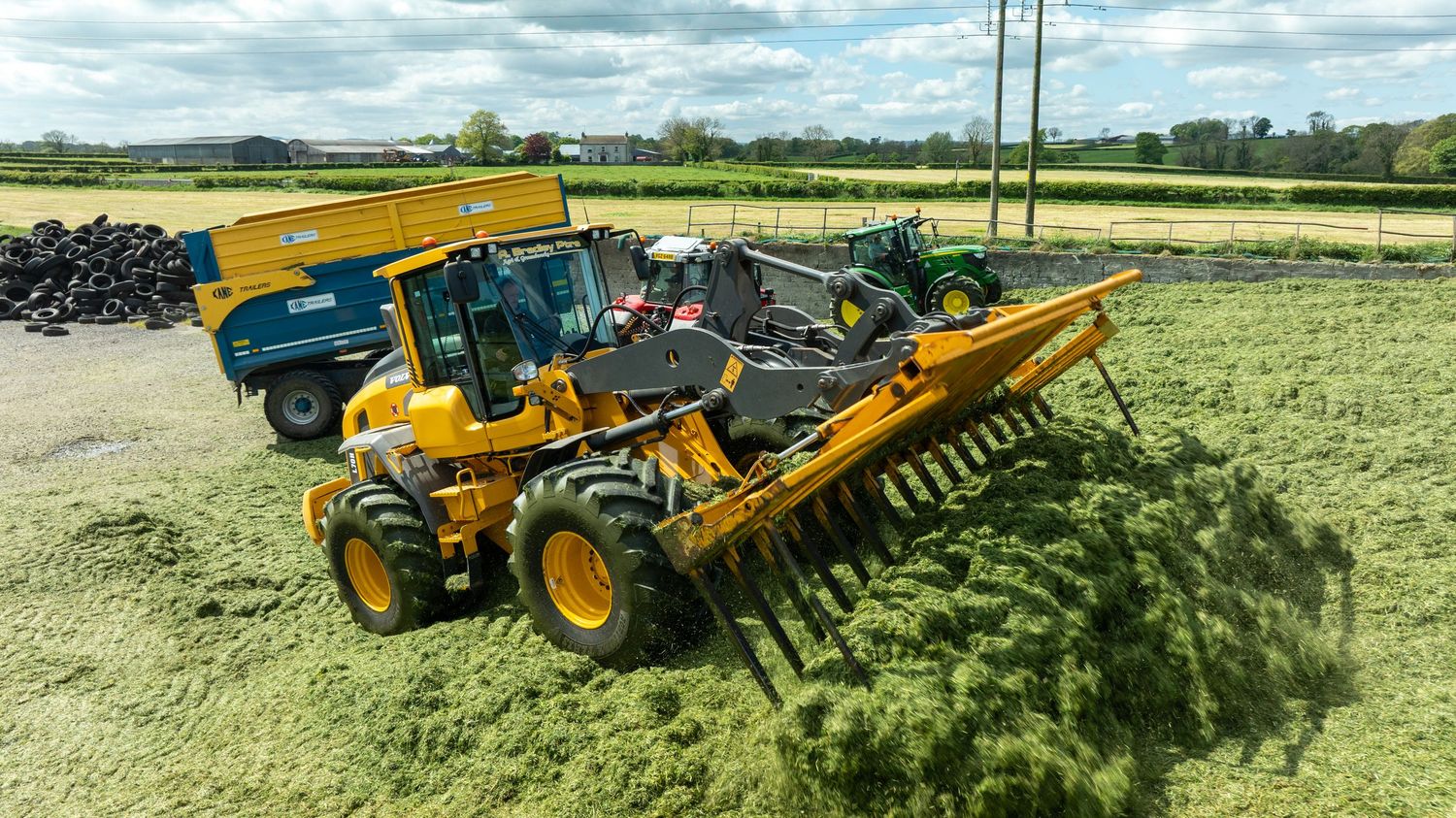 Yellow construction vehicle with large rake attachment spreading green materials across an agricultural field.