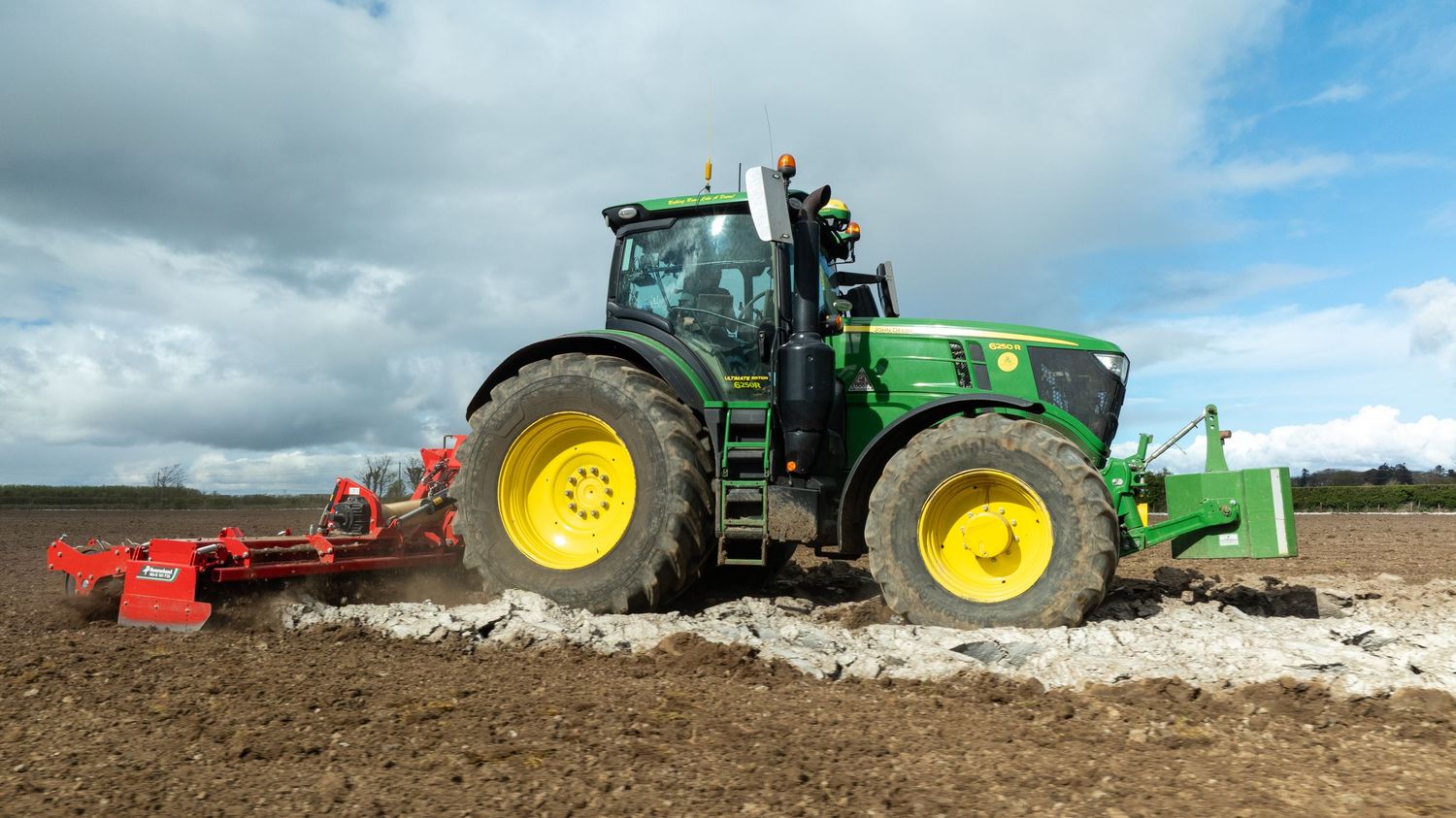 Green John Deere tractor tilling brown soil with farming equipment on cloudy day in agricultural field.