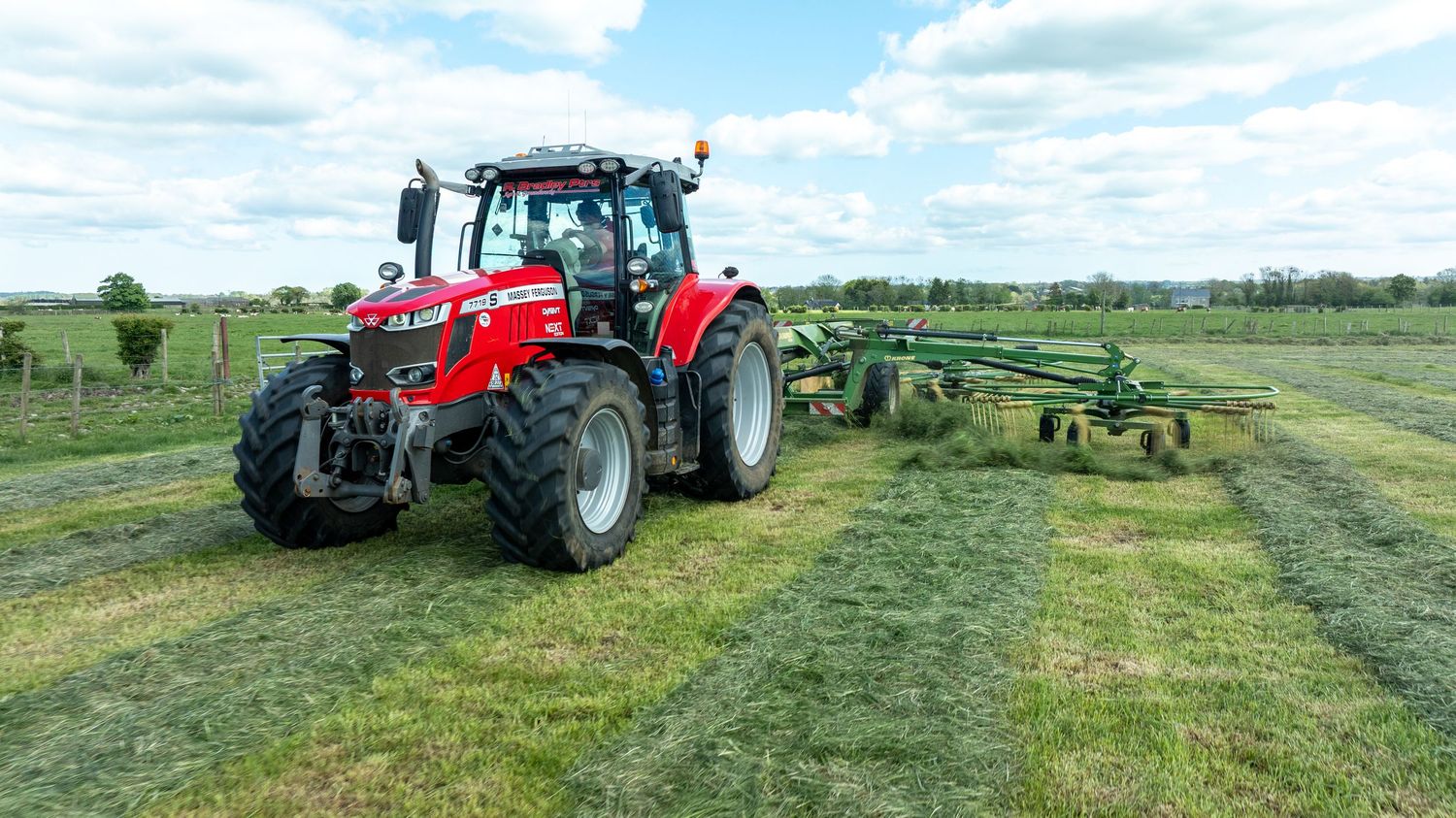 Red Massey Ferguson tractor with hay raking equipment working in green agricultural field under cloudy sky.