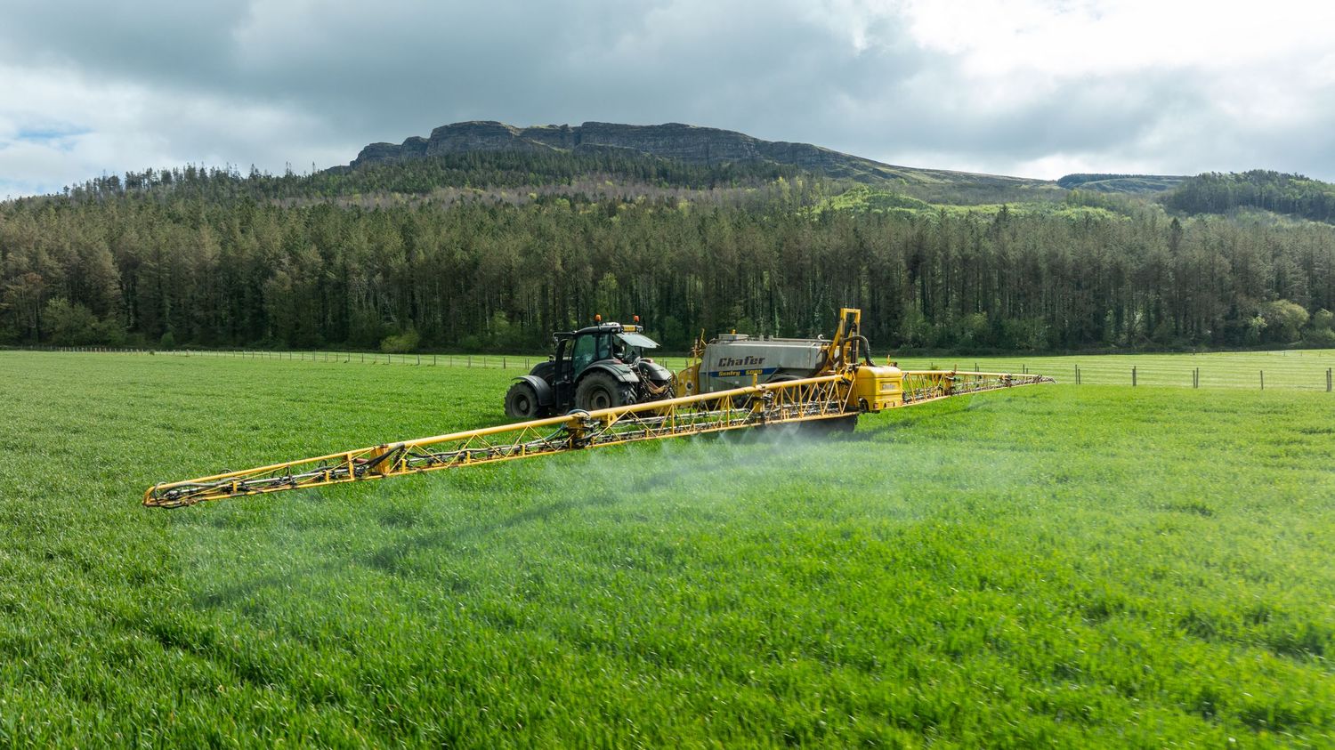 Agricultural tractor spraying pesticides with wide boom sprayer on green grass field in hilly landscape.