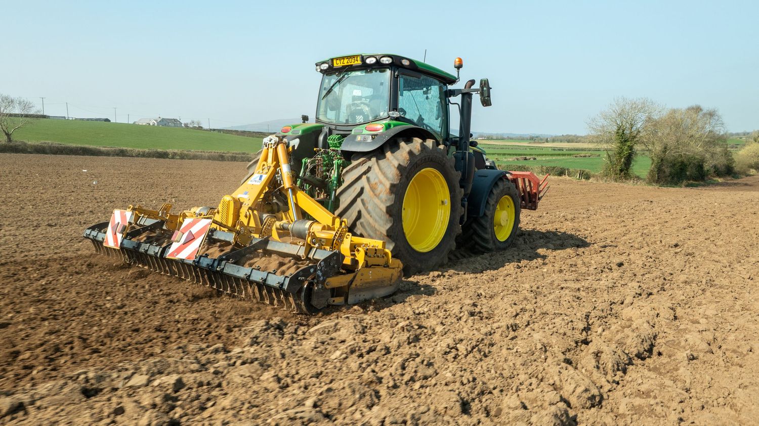 Modern green John Deere tractor with tillage equipment working in a brown agricultural field on a sunny day.
