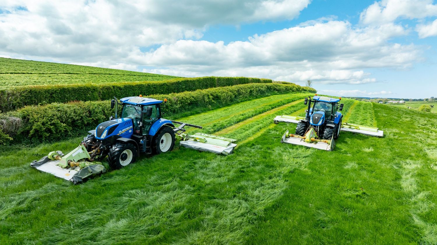 Two blue tractors with mowing attachments cutting grass in parallel on a lush green hillside field under cloudy skies.