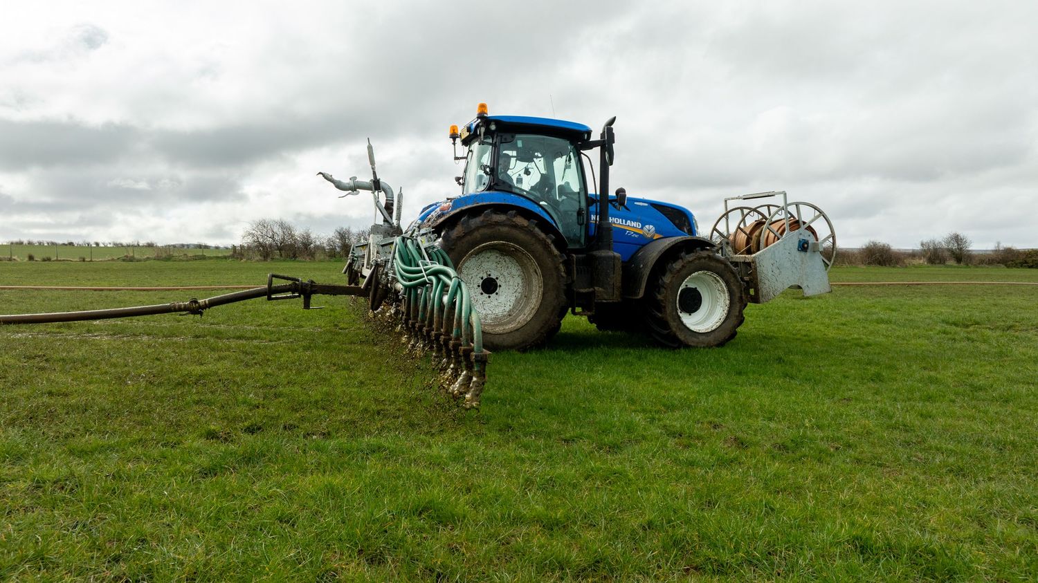 A blue tractor with mounted sprayer equipment working in a green pasture field under cloudy skies.