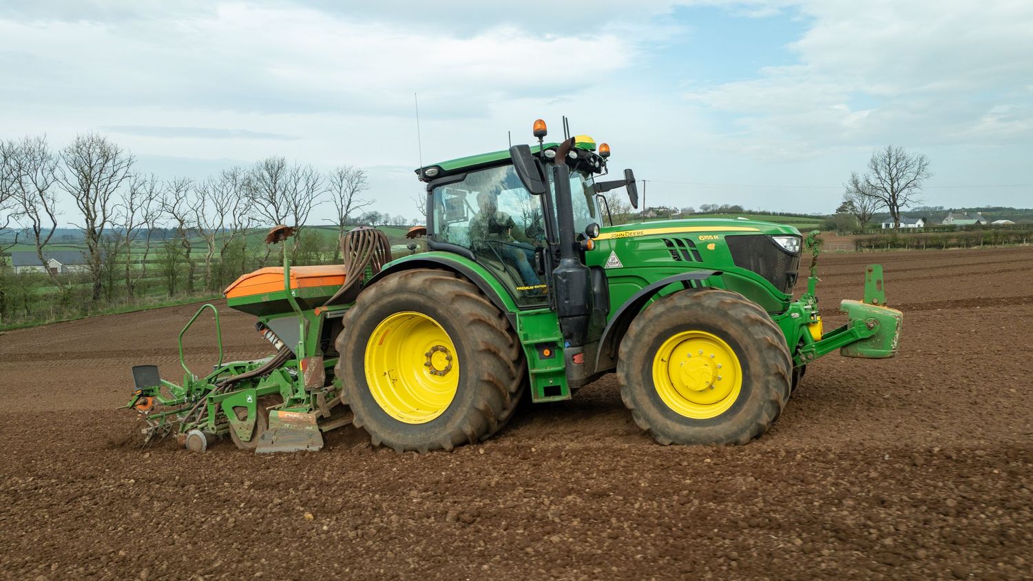 Green John Deere tractor plowing rich brown soil in a farm field on a partly cloudy day.