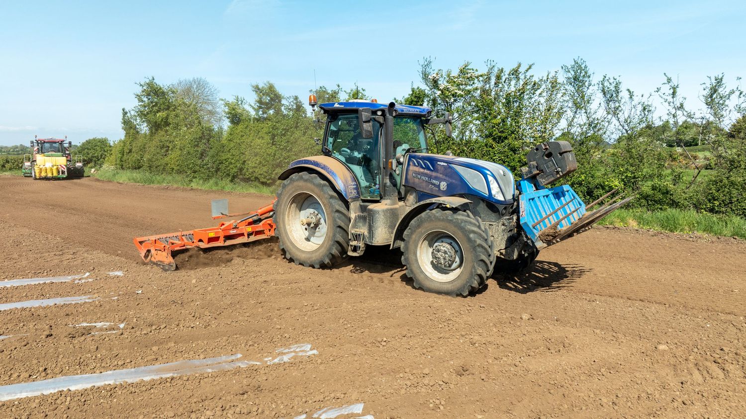 Blue New Holland tractor with cultivator attachment preparing soil in agricultural field on sunny day.