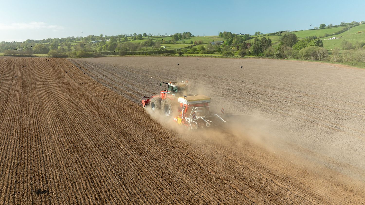 A tractor towing farming equipment kicks up dust while working in a large brown plowed field on rolling hills.