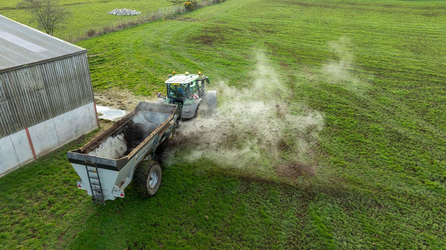 Aerial view of a tractor spreading fertilizer on green farmland next to a storage building.
