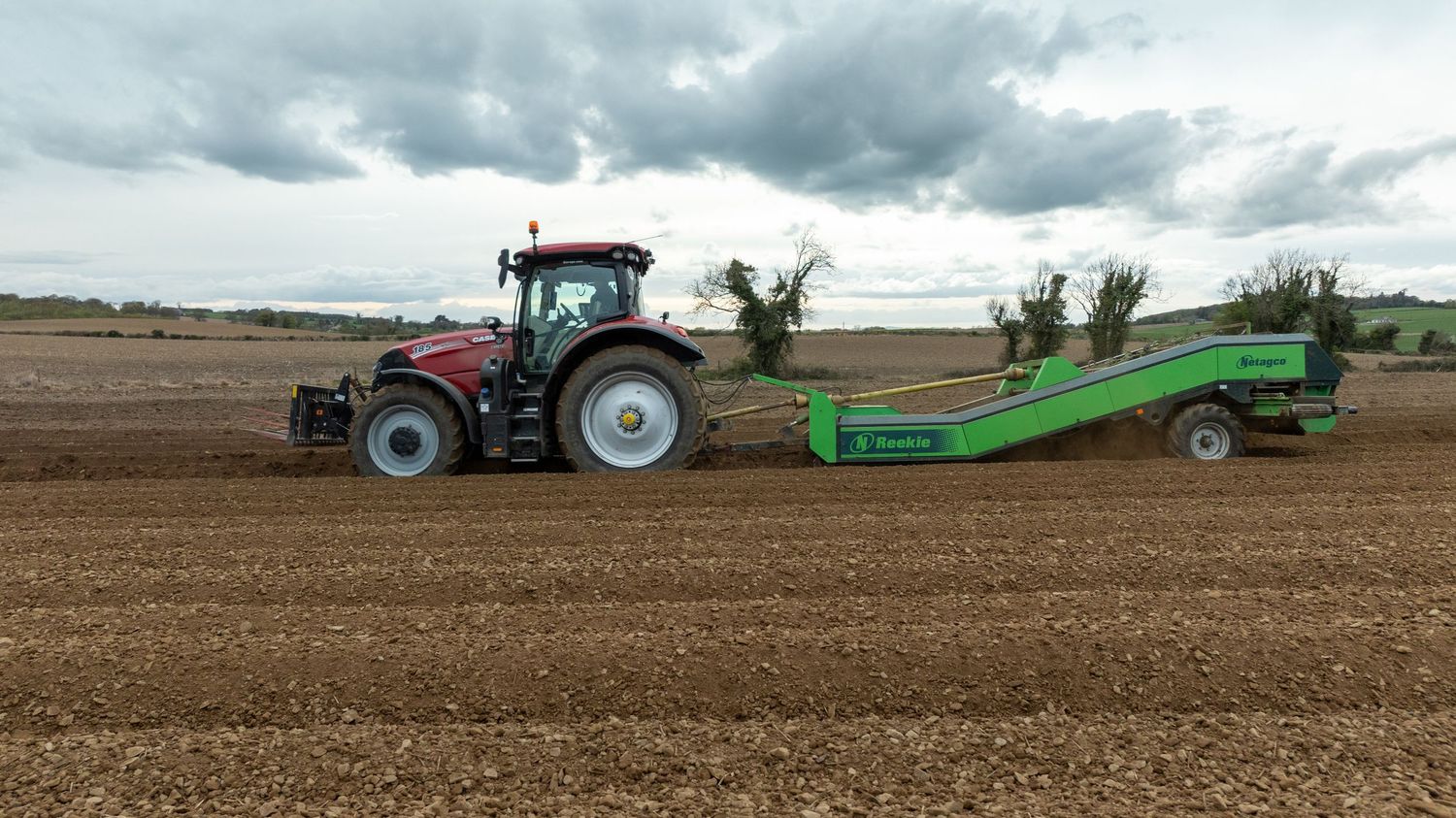 Red tractor pulling a green agricultural implement across a freshly plowed field under dramatic cloudy skies.