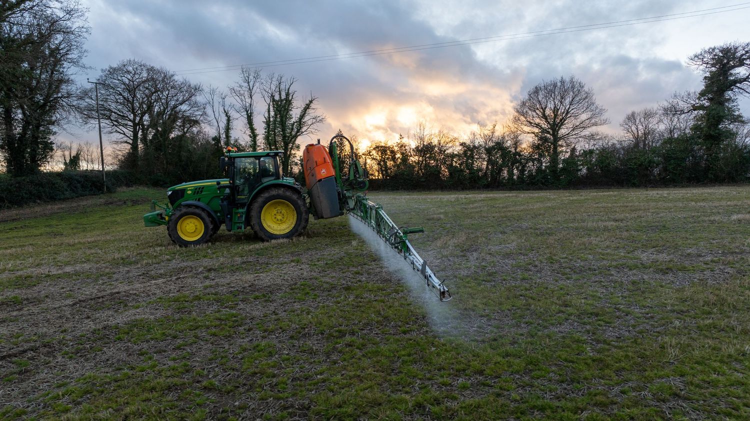 Agricultural tractor spraying fertilizer on a field at sunset with dramatic sky and silhouetted trees in background.