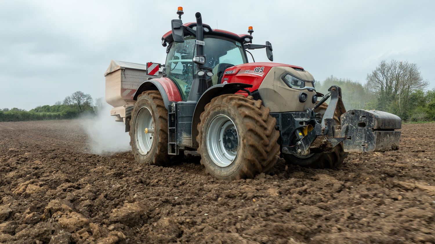 Red modern tractor spraying and cultivating farm field soil with large tires in overcast conditions.