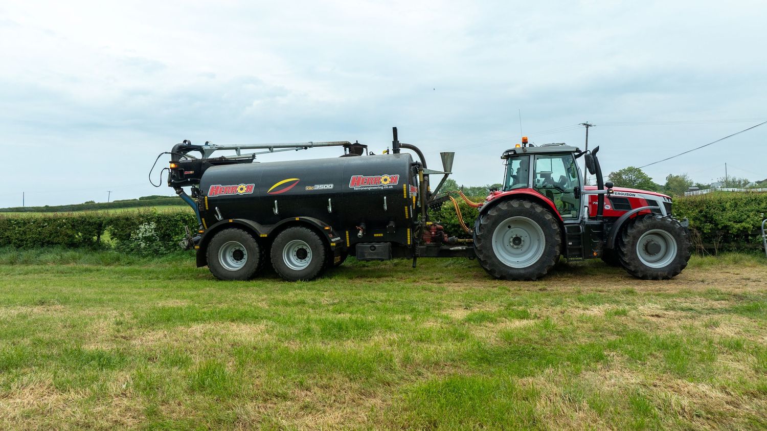 Red tractor pulling a large slurry tanker on green farmland under cloudy skies.