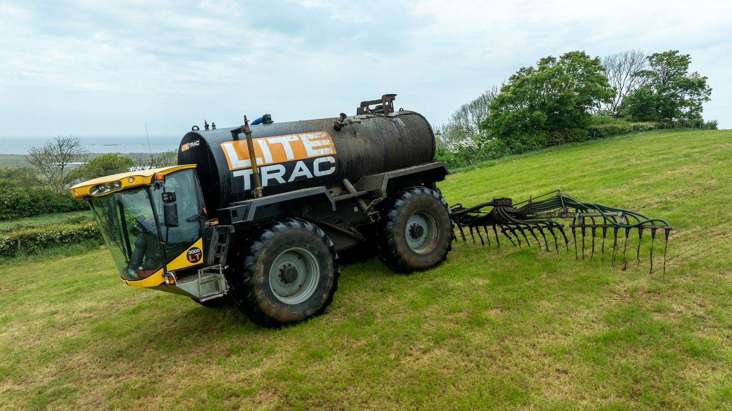 Large agricultural sprayer tanker with yellow cab parked on green hillside with drag bar implements attached.
