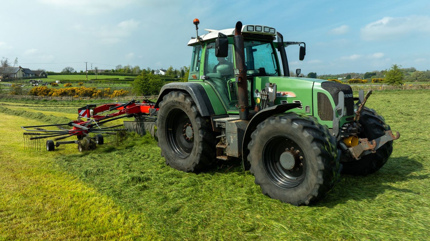 Green agricultural tractor with cultivation equipment parked on grassy field under blue sky.