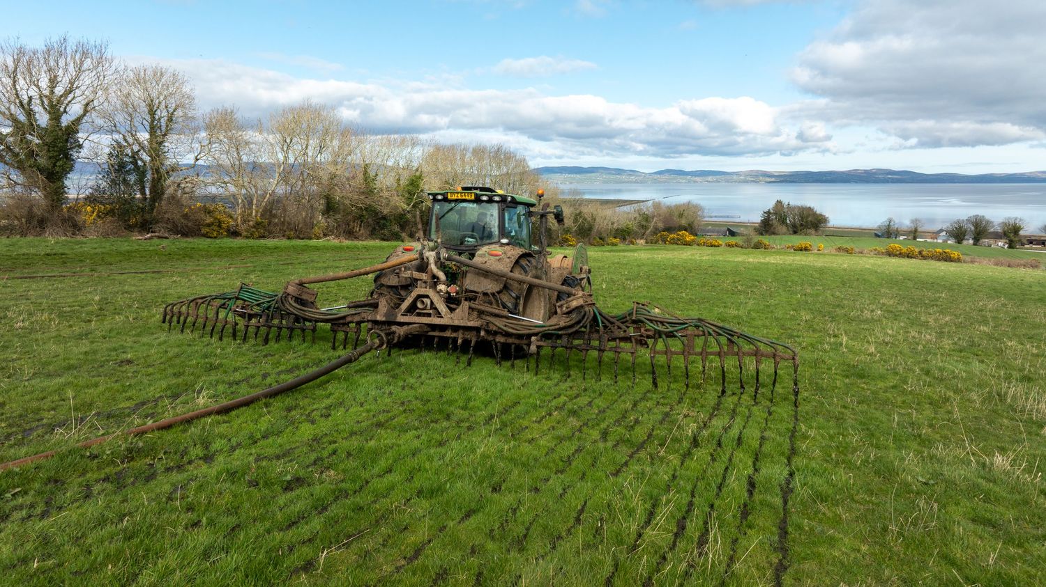 A tractor pulling a large harrow implement across a green pasture field with trees in the background.