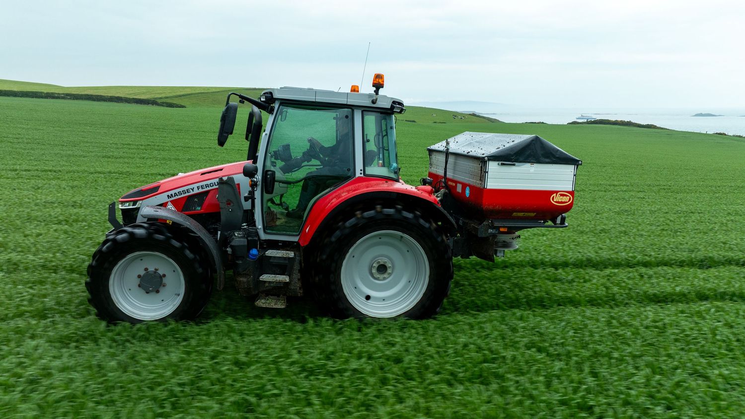 Red tractor with fertilizer spreader attachment operating in a lush green agricultural field.