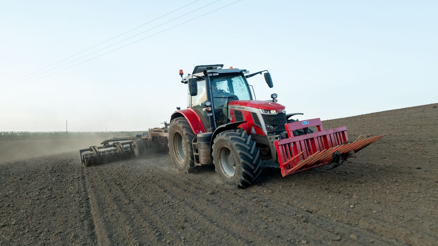 Red tractor with plowing equipment working in a dusty farm field on a hazy day.