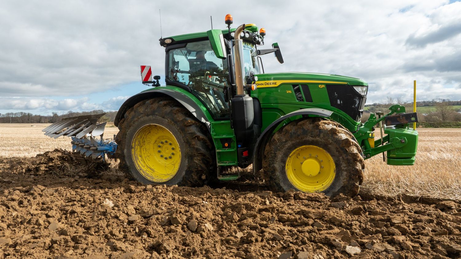 A green John Deere tractor plows through brown soil in an agricultural field under cloudy skies.