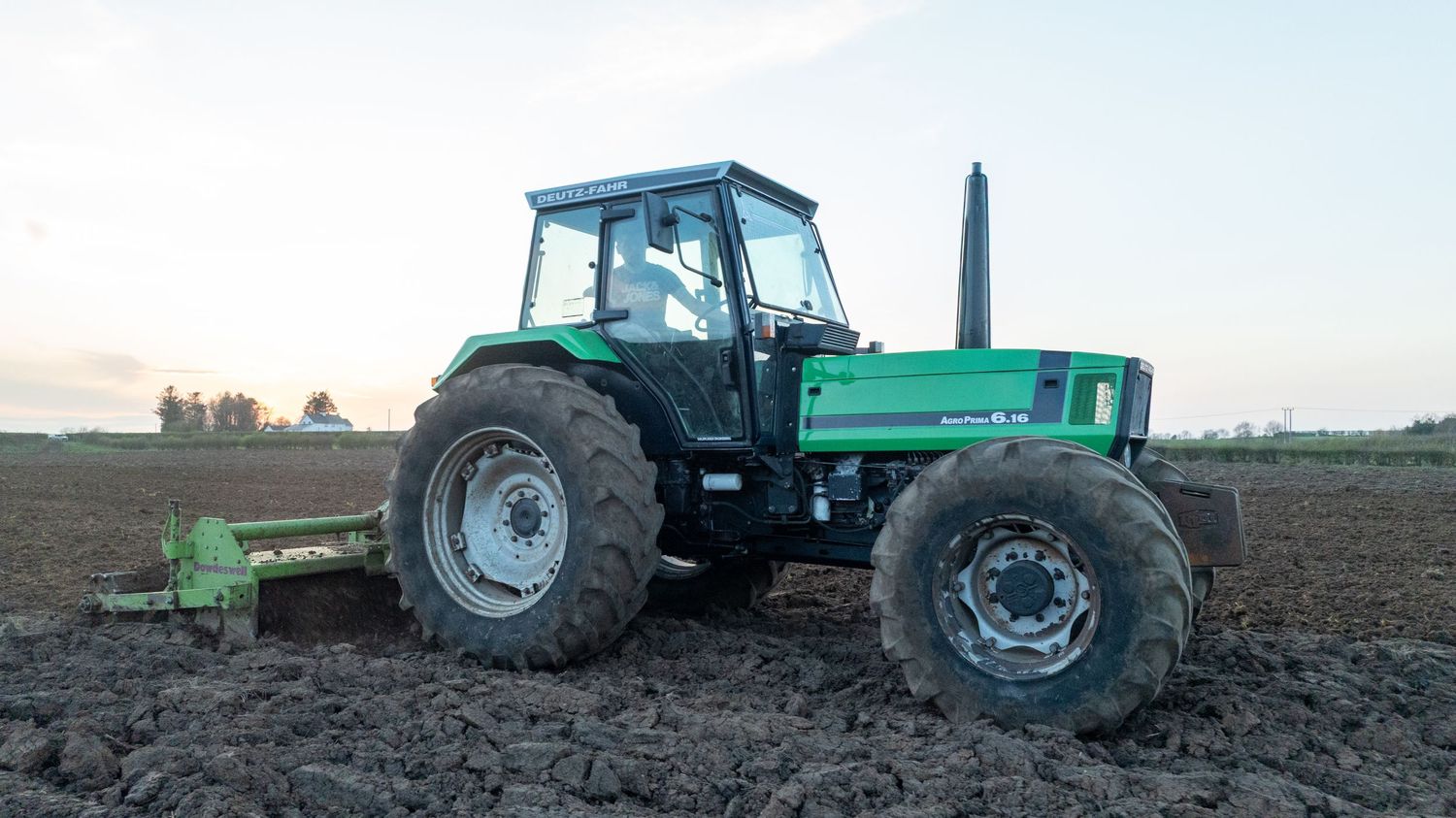 Green tractor with a plowing implement works in a muddy field during early evening hours.