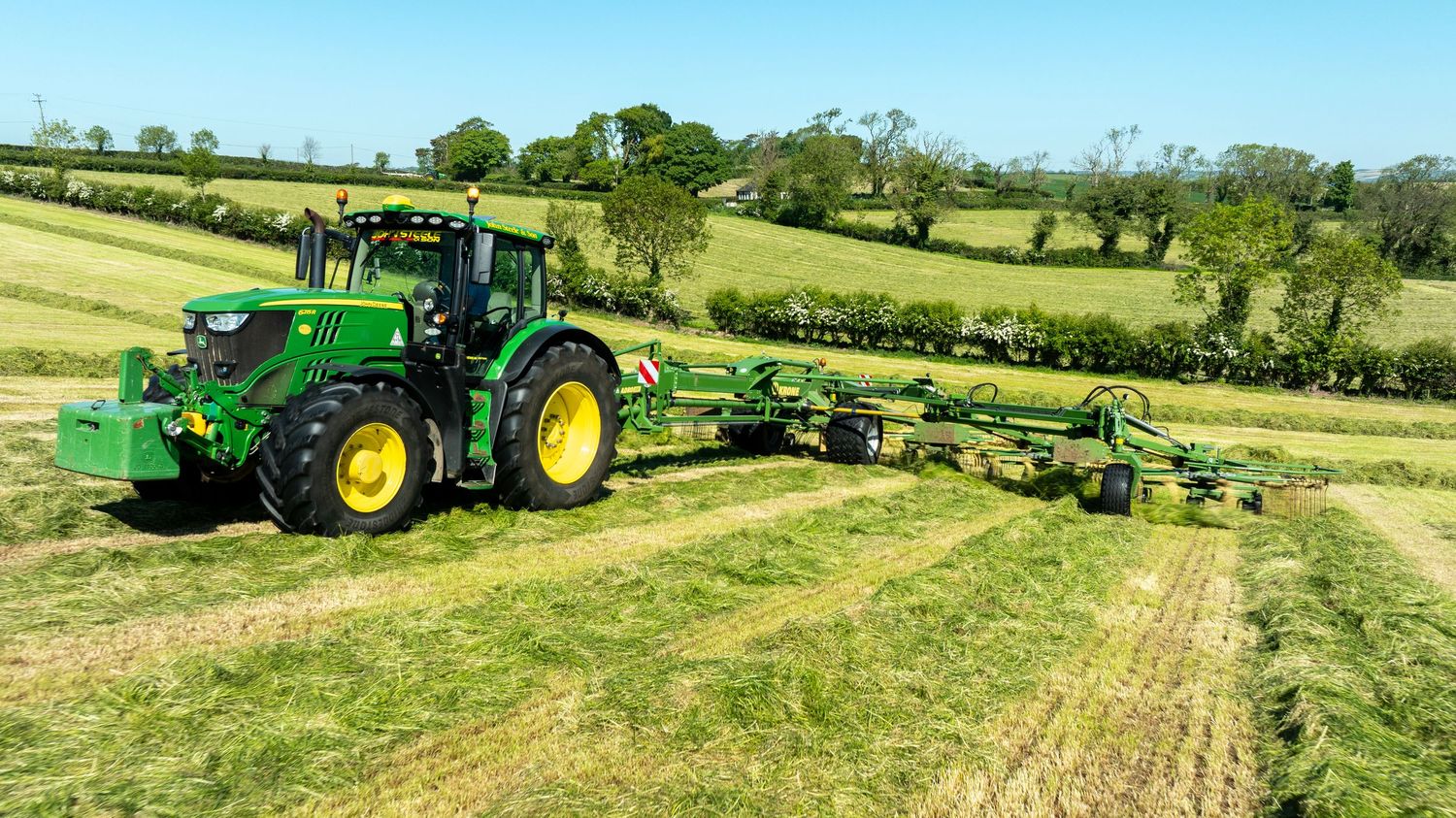 John Deere tractor with triple mower attachment cutting grass hay in a rolling green countryside field.