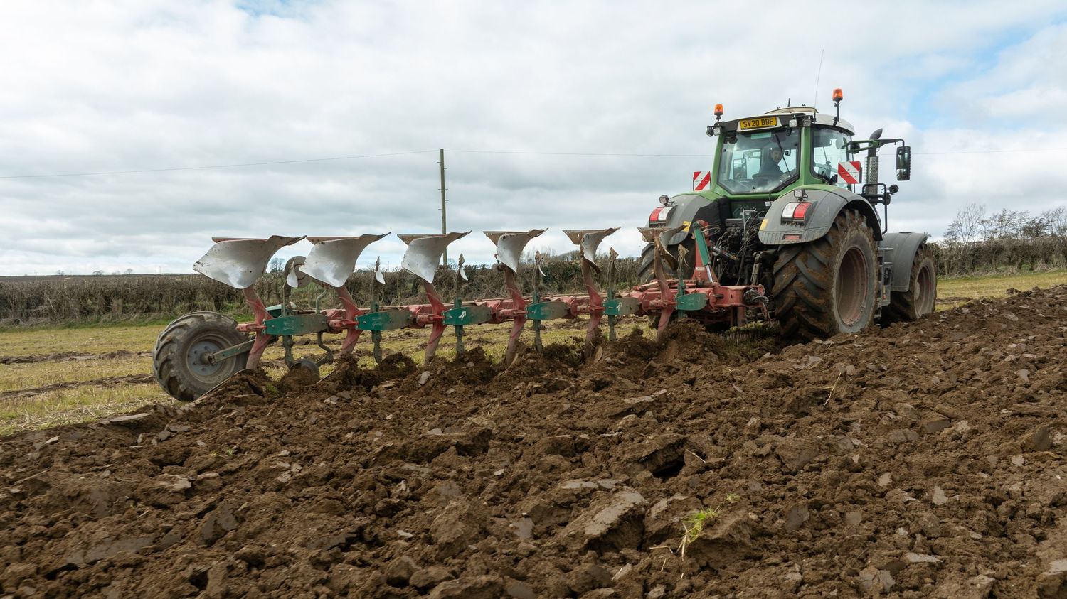 A modern tractor pulls a large multi-blade plow through brown soil in an agricultural field under cloudy skies.