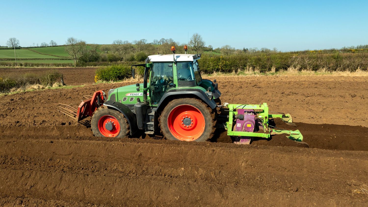Green Fendt tractor with red-rimmed wheels plowing brown soil in a rural agricultural field on a sunny day.