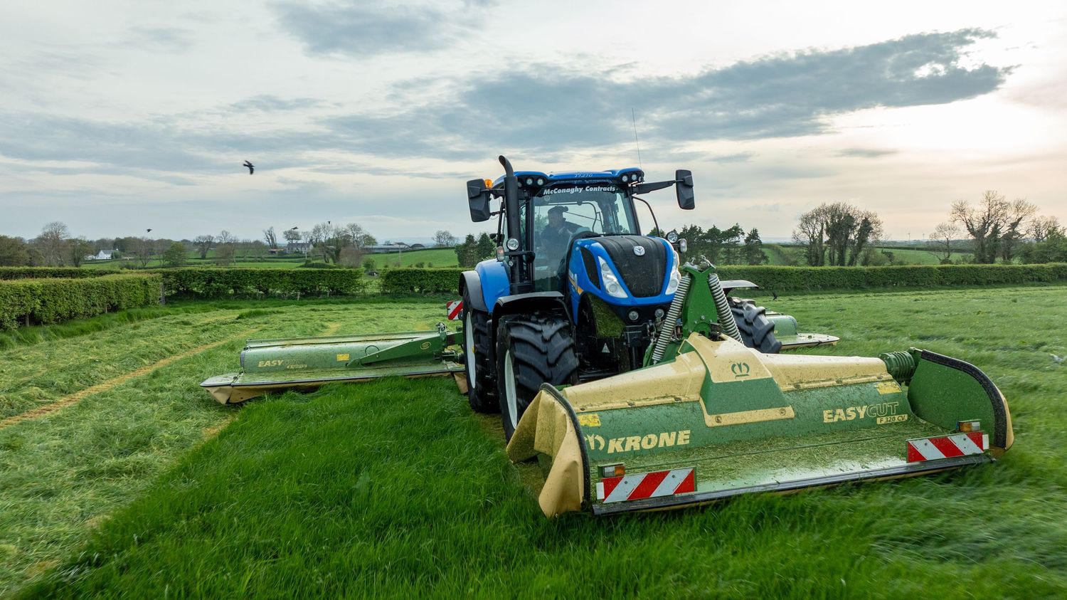 A blue tractor mowing a green field with a large cutting attachment at sunset in rural farmland.