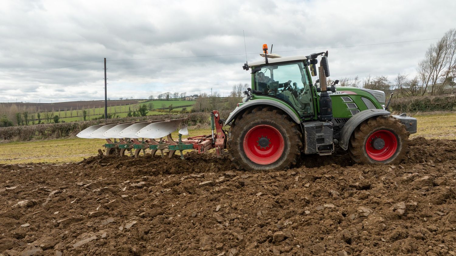 Green Fendt tractor with large plow attachment tilling dark soil in a rural field under cloudy skies.