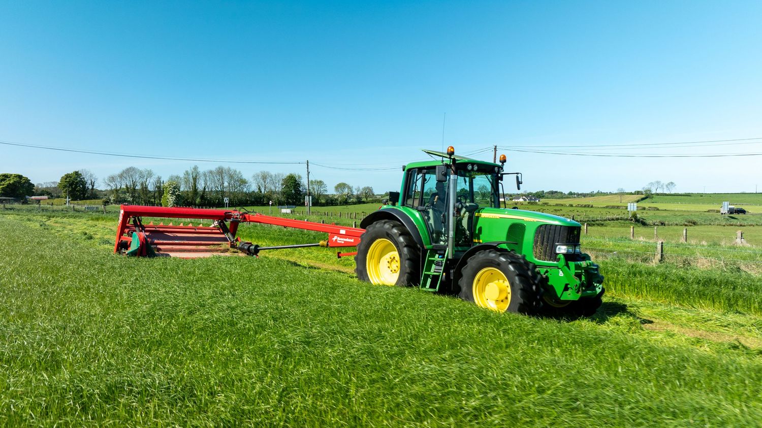 Green John Deere tractor pulling red hay equipment across a sunny farm field on a clear summer day.