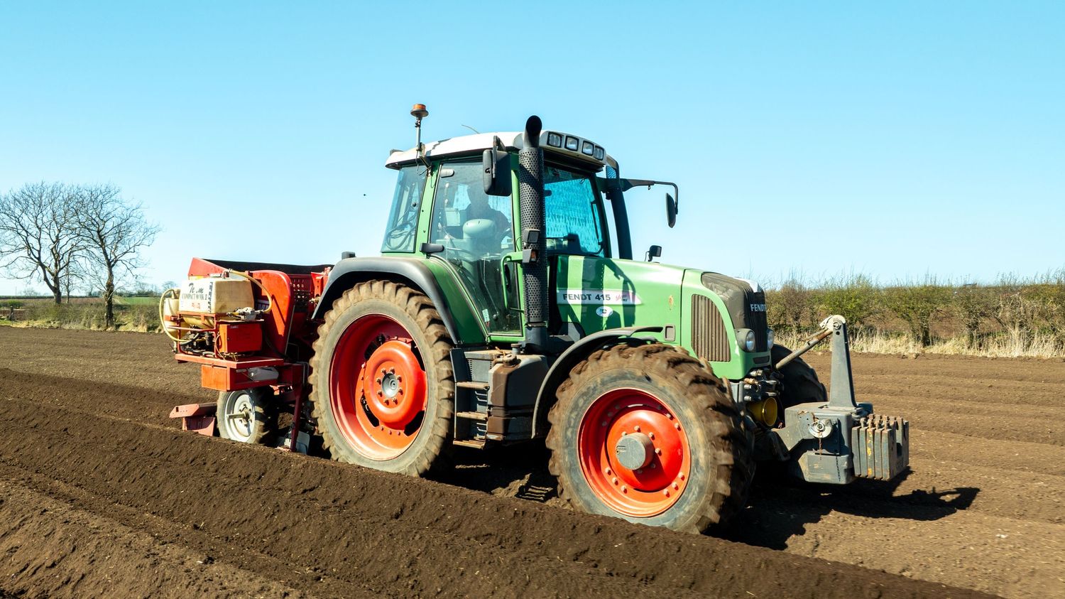 A green Fendt tractor with red wheels plowing a brown field on a sunny day with blue sky in the background.