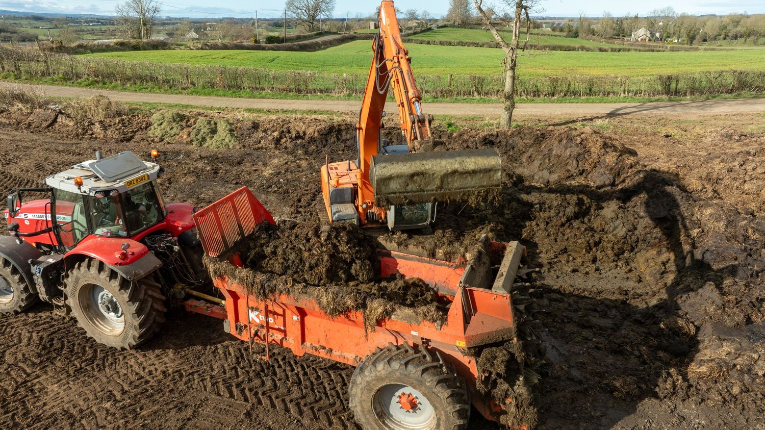 Orange excavator and red tractor working together to move soil on a construction site in rural countryside.