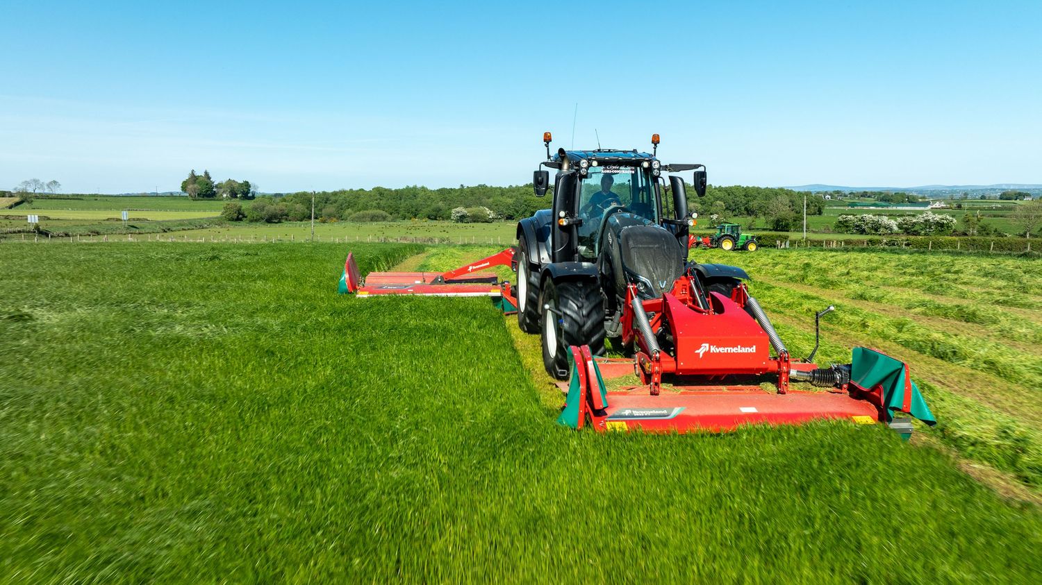 Tractor pulling red mowing equipment across lush green pasture field under blue sky.