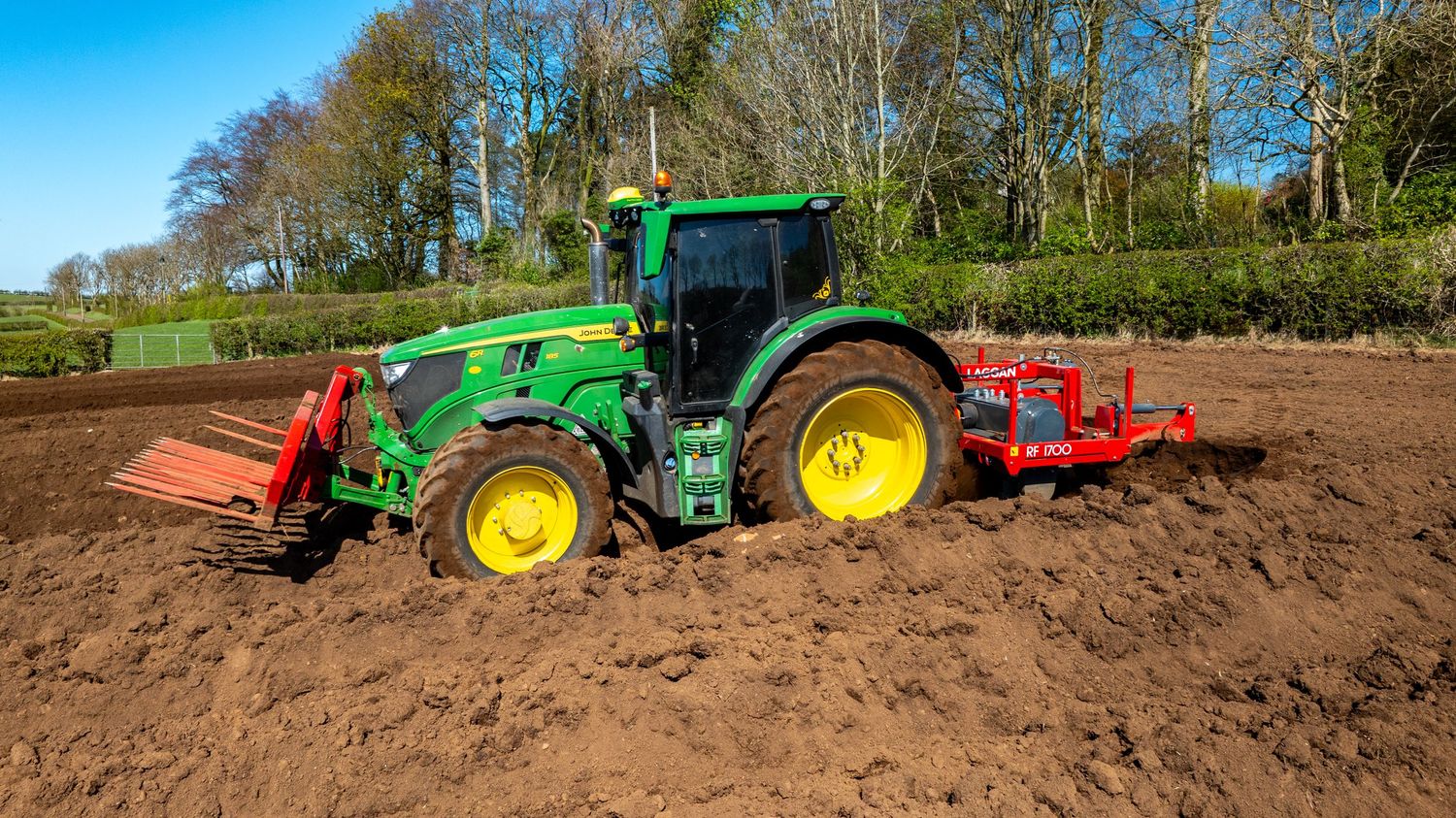 John Deere tractor with front loader attachment cultivating dark soil near a tree line on farmland.
