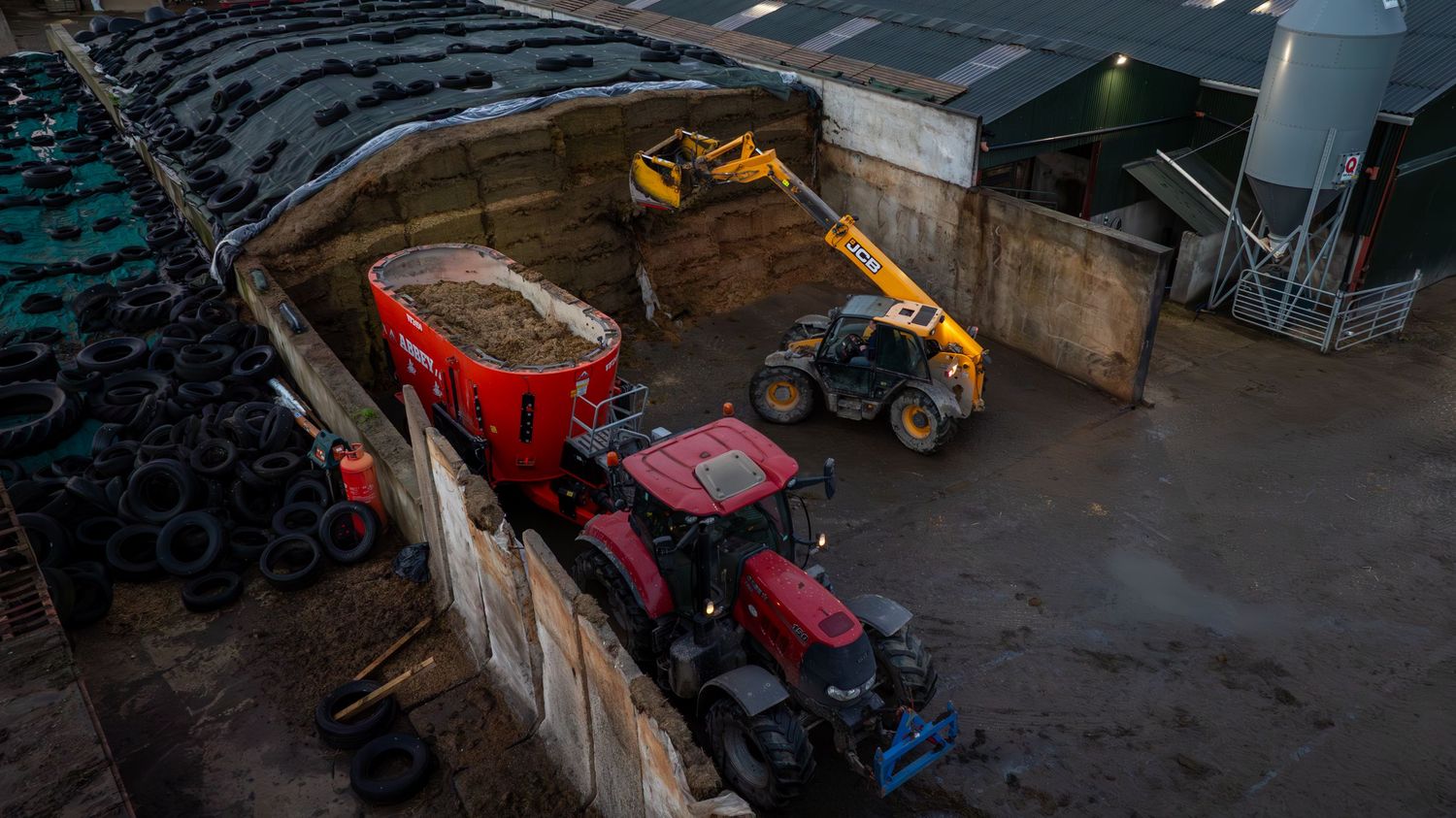 Aerial view of red tractor and loader machinery working together at agricultural storage facility at dusk.
