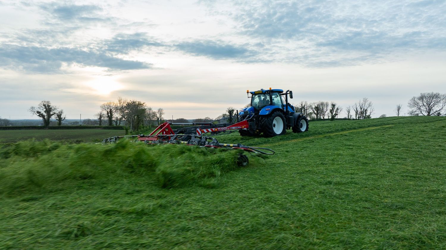 Blue tractor with farm equipment working on a green hillside field during sunset with cloudy sky.
