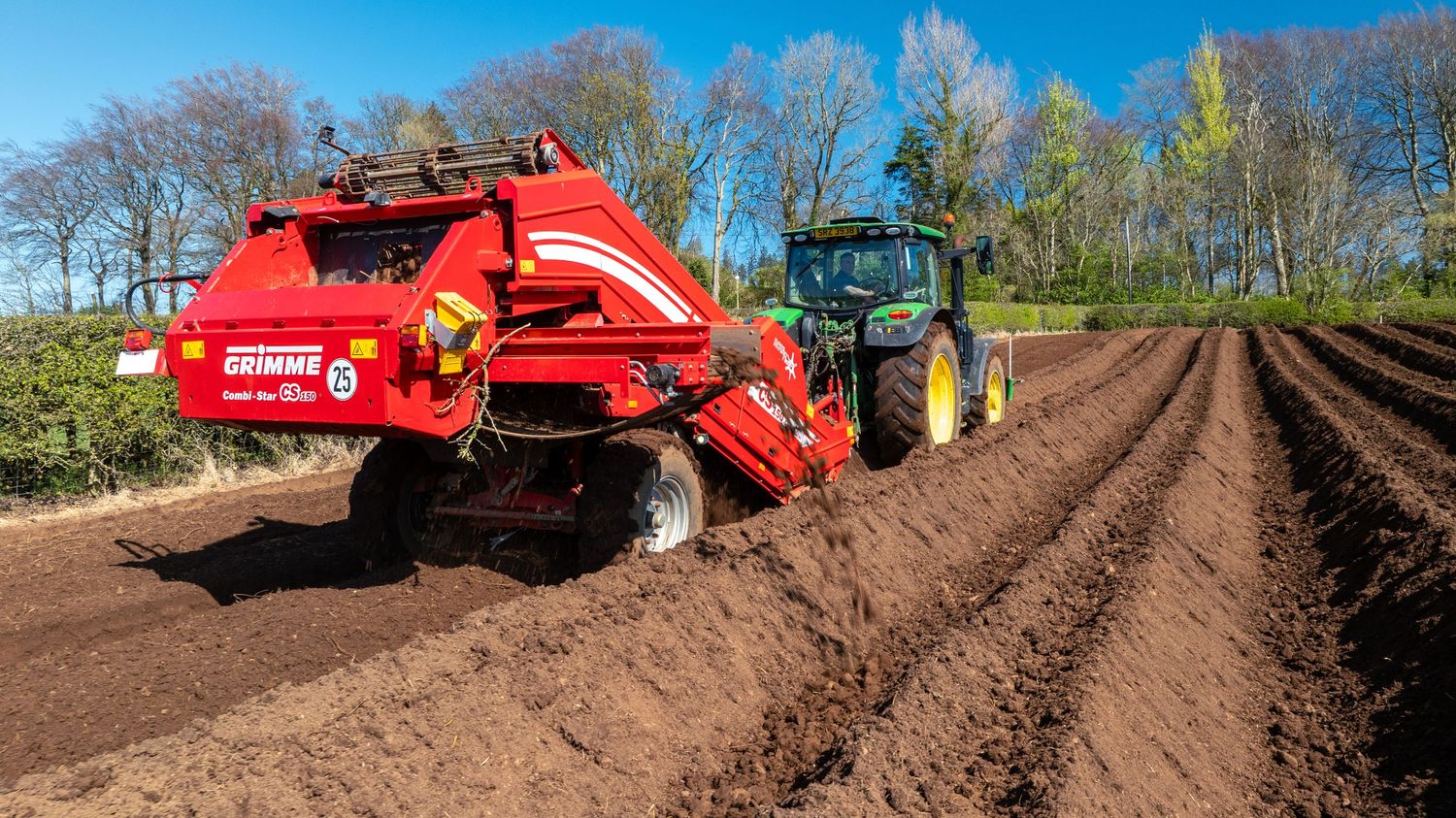 Green tractor pulling red farming equipment creating neat rows in freshly tilled brown soil on a sunny spring day.