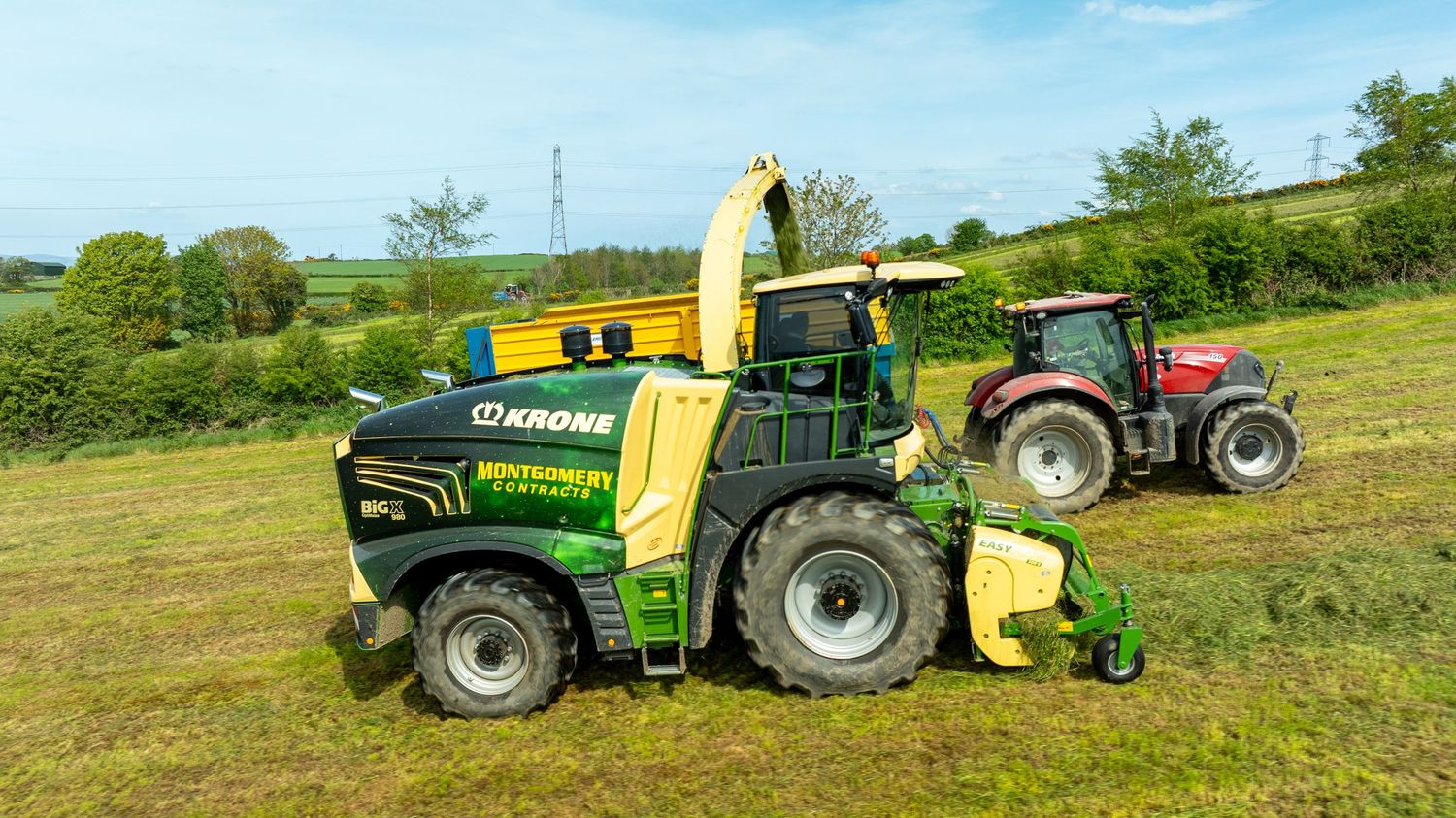 Green forage harvester working in a grassy field alongside a red tractor on rolling farmland.