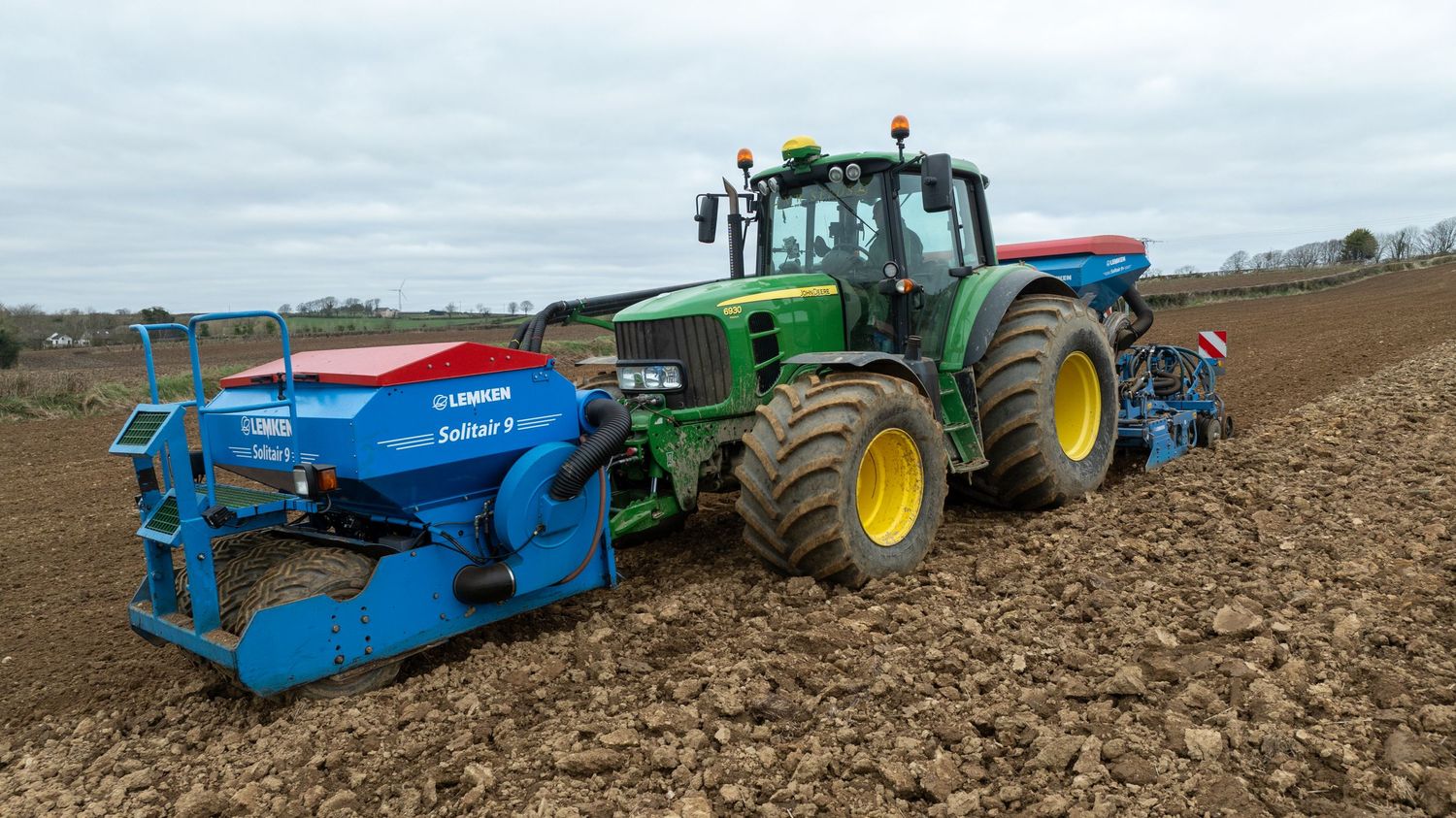 A green John Deere tractor with blue agricultural equipment attachment plows through a brown field on an overcast day.