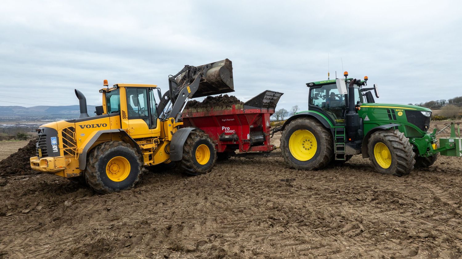 Heavy machinery including a yellow front loader and green tractor work together loading soil on agricultural field.