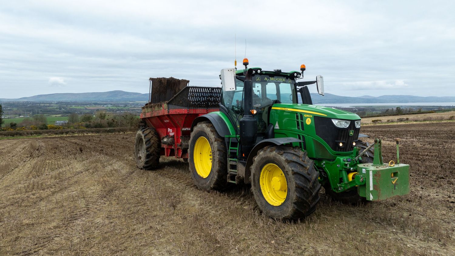 Large green John Deere tractor with red fertilizer spreader attachment working in an agricultural field on overcast day.