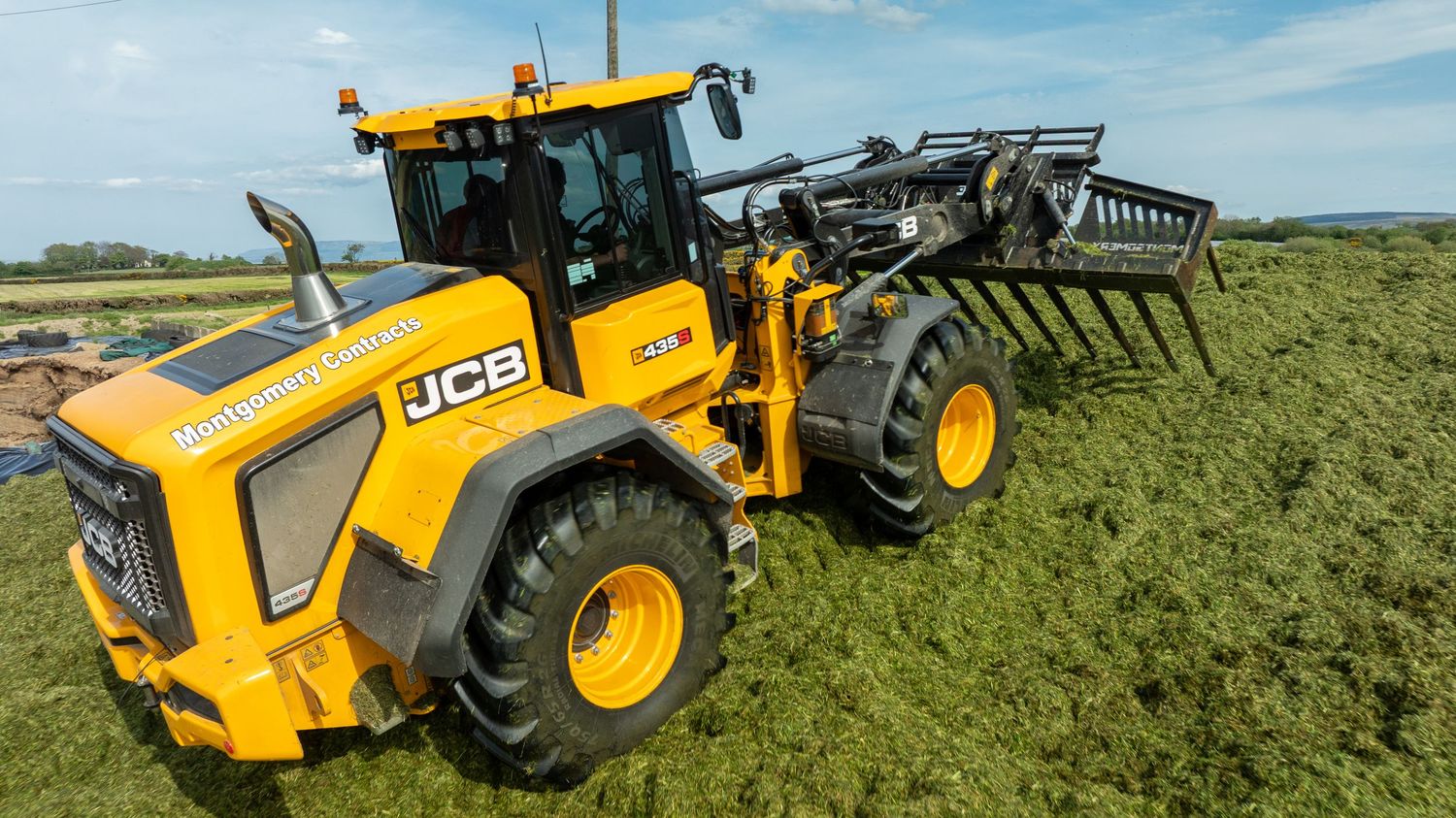 Yellow JCB wheel loader working on silage pile at dairy farm with blue sky in background.
