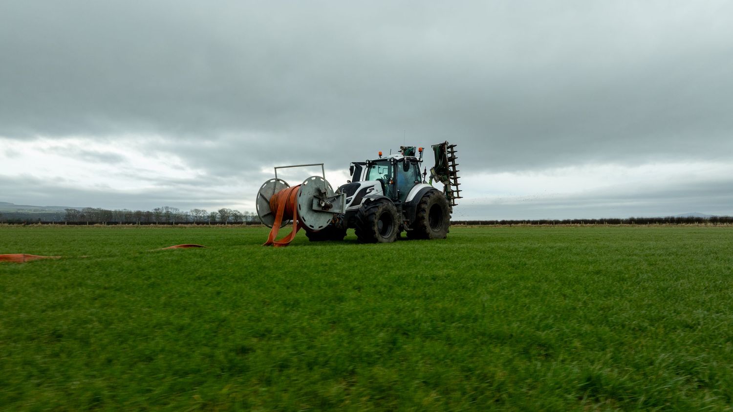 A heavy-duty backhoe loader sits in a vast green field under overcast skies during agricultural work.
