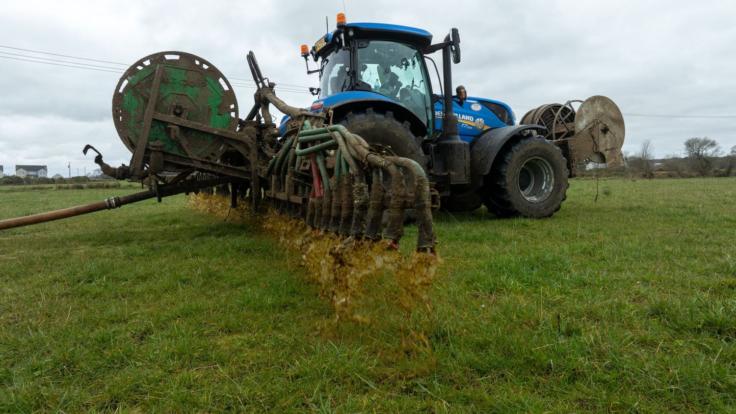 Blue tractor with manure spreader equipment operating in a green field under cloudy skies.