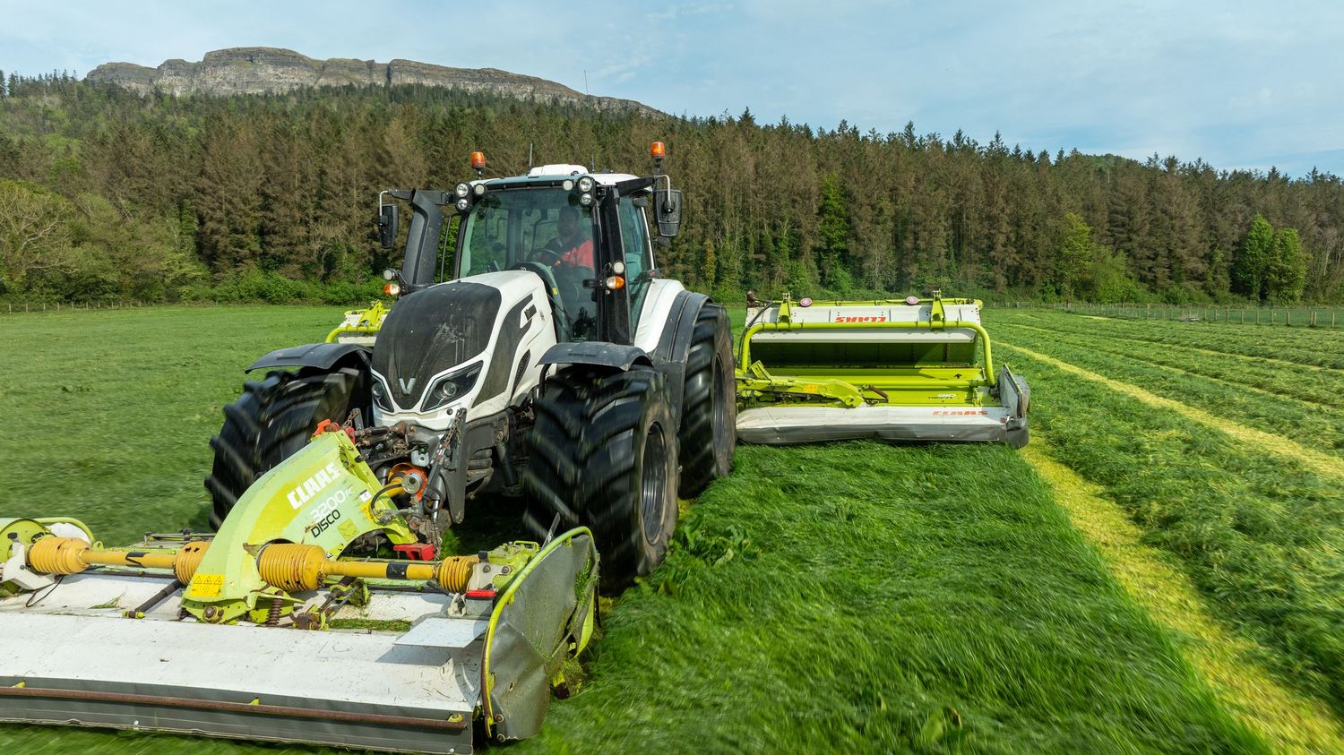Modern tractor with forage harvesting equipment cutting grass in a lush green field near forest hills.
