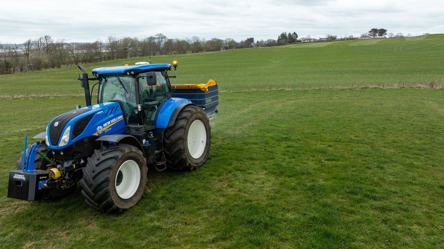 A blue New Holland tractor parked on lush green farmland under an overcast sky.